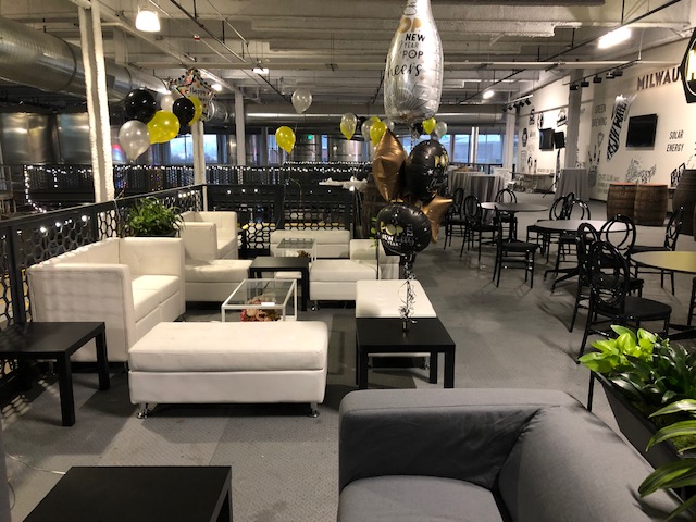 A modern lounge area decorated with black, gold, and silver balloons—including a champagne bottle balloon—offers stylish holiday party ideas. White sofas, black tables, greenery, and string lights enhance the festive atmosphere.