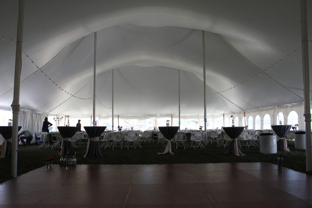 Large white event tent with round tables and chairs arranged inside, cocktail tables covered in black cloths, string lights overhead, and a few people setting up the venue—perfect for those seeking creative holiday party ideas. The floor in front appears to be a dance area.