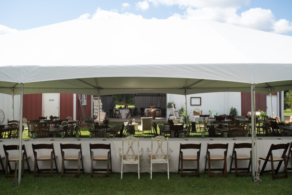 A large white outdoor tent with various wooden chairs and tables arranged underneath, perfect for holiday party ideas, set up on green grass. The background features a barn and clear blue sky with clouds.
