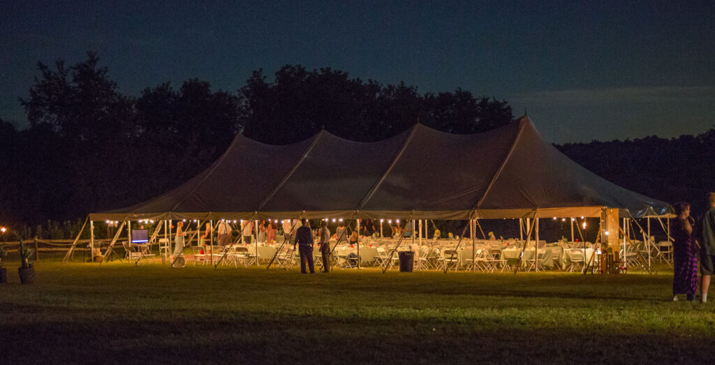 A large outdoor tent is set up at night, warmly lit inside with string lights and decorated tables—a perfect setting for gathering and sharing holiday party ideas. A few people stand and converse near the entrance on a grassy field surrounded by trees.