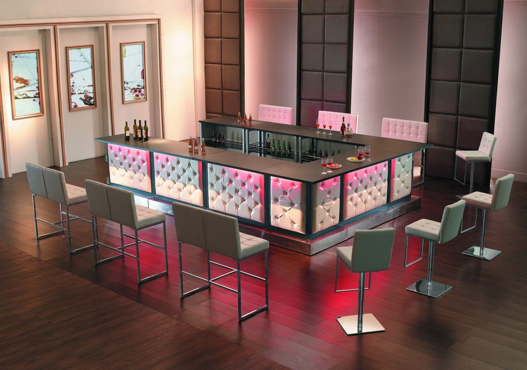 A modern bar with white padded panels, illuminated by pink lights, stands in the center of a room—perfect for holiday party ideas. Tall chairs surround the bar, and bottles and glasses are arranged on the dark wood floor.