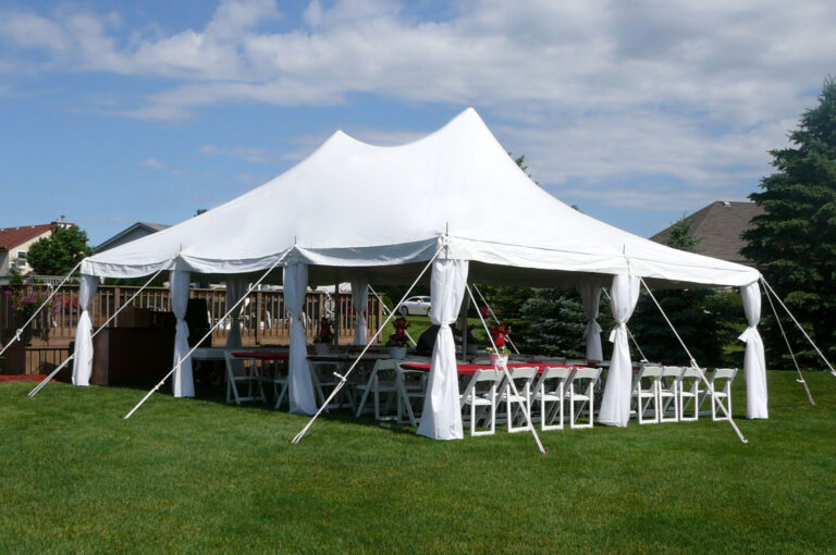 A large white event tent is set up on a grassy lawn, with tables and chairs arranged underneath—perfect for holiday party ideas. Supported by poles and ropes, the tent sits beneath a partly cloudy sky with trees and houses in the background.