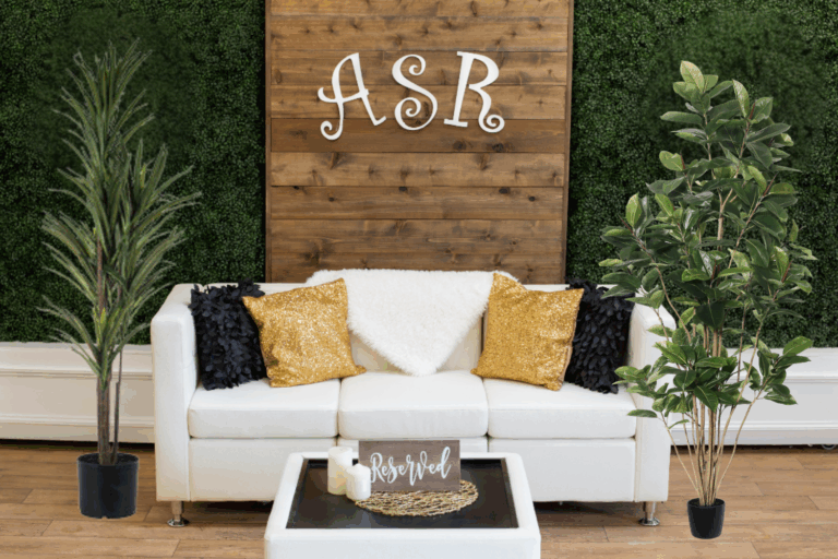A white sofa with gold and black pillows sits in front of a wooden wall with the letters ASR—perfect holiday party ideas inspiration. Potted plants flank the sofa, and a table with candles and a Reserved sign is in front. Greenery covers the background wall.