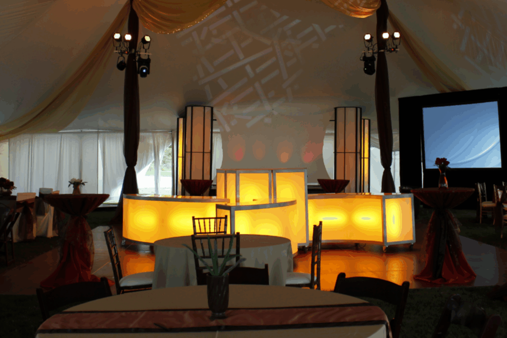 A decorated event tent with draped ceilings, a glowing yellow bar at the center, round tables with beige tablecloths, chairs, and a large projection screen in the background—perfect for inspiring holiday party ideas.