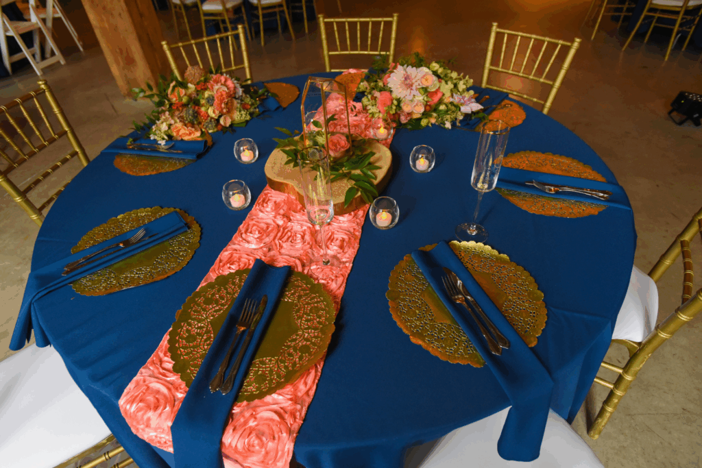 A round table with a blue tablecloth, gold chairs, gold placemats, navy blue napkins, and a pink floral runner—perfect for holiday party ideas. The centerpiece features flowers, greenery, a glass lantern, and small candles.