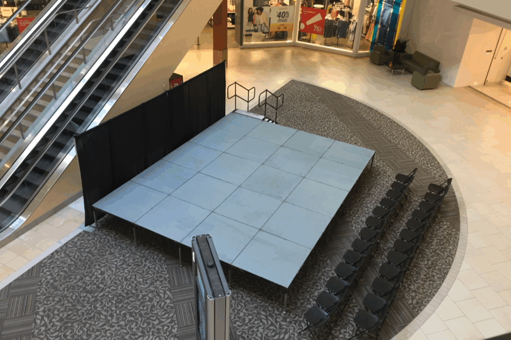 A small empty stage with steps and black backdrop is set up in a mall near an escalator, with rows of black chairs arranged in front—an inviting spot for showcasing holiday party ideas. Storefronts and mall seating are visible in the background.