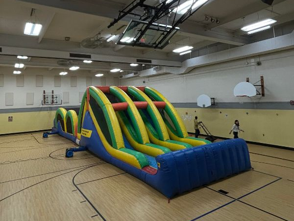A large inflatable obstacle course is set up in the middle of a gymnasium with wooden floors and basketball hoops—an exciting option for holiday party ideas. A person stands nearby as ceiling lights illuminate the lively space.