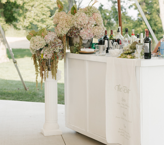 A white bar setup outdoors under a tent, decorated with large floral arrangements and stocked with wine, spirits, glassware, and fresh grapes—an inspiring scene for memorable holiday party ideas. A sign with drink options hangs on the front of the bar.
