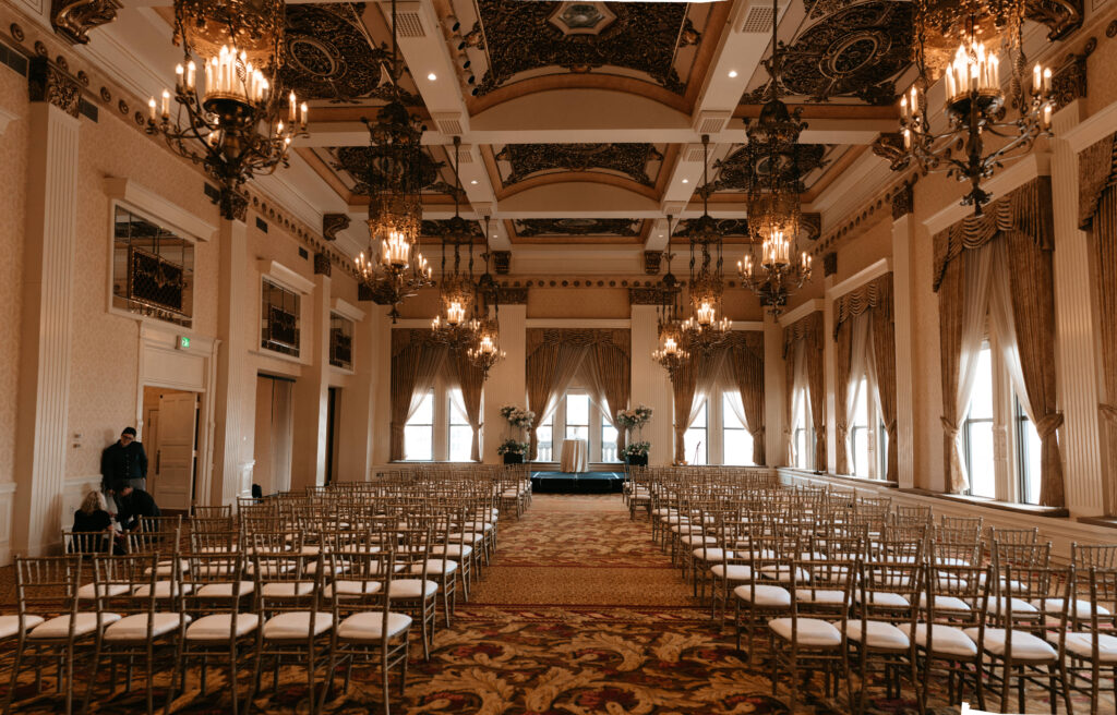 A grand, elegant ballroom with ornate chandeliers, high decorated ceilings, and rows of gold chairs facing a small stage—perfect for sophisticated holiday party ideas. Large windows with draped curtains let in natural light. Two people stand near the left wall.