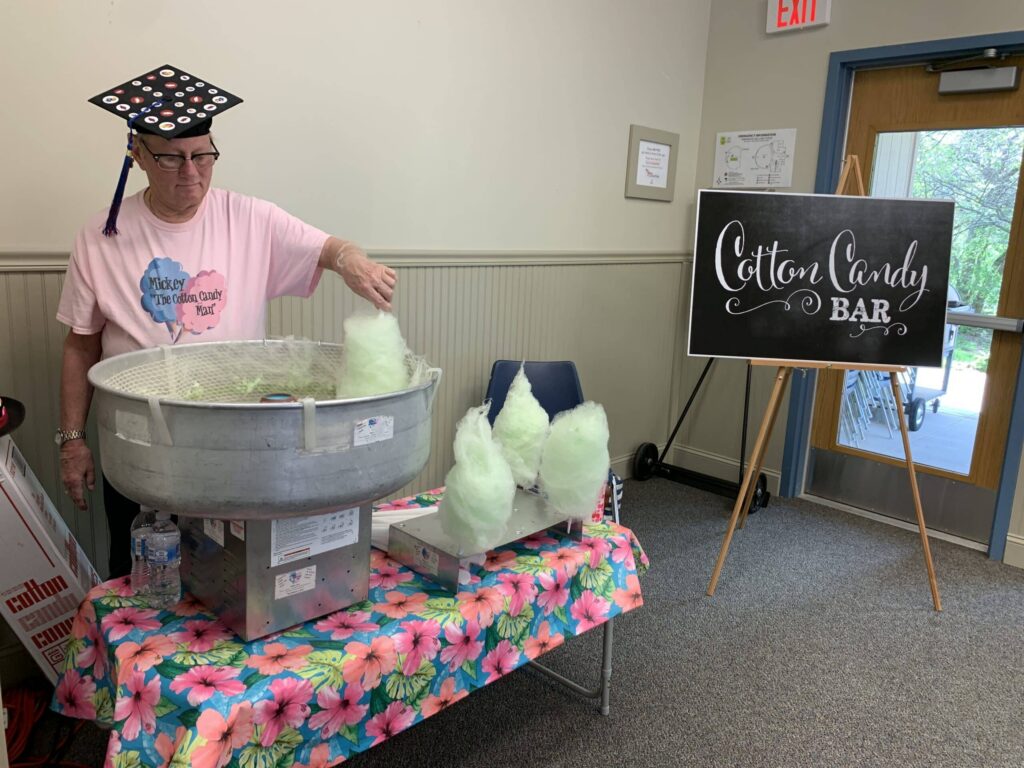 A person wearing a decorated graduation cap and a pink t-shirt makes cotton candy at a table with several finished cones—perfect for graduation party rentals. A chalkboard sign reads Cotton Candy Bar atop a colorful floral cloth.