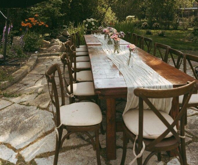 A long wooden outdoor table with cushioned chairs and a white runner, set with small vases of pink flowers, showcases 2026 wedding rental trends amid trees and garden plants under a large black umbrella.