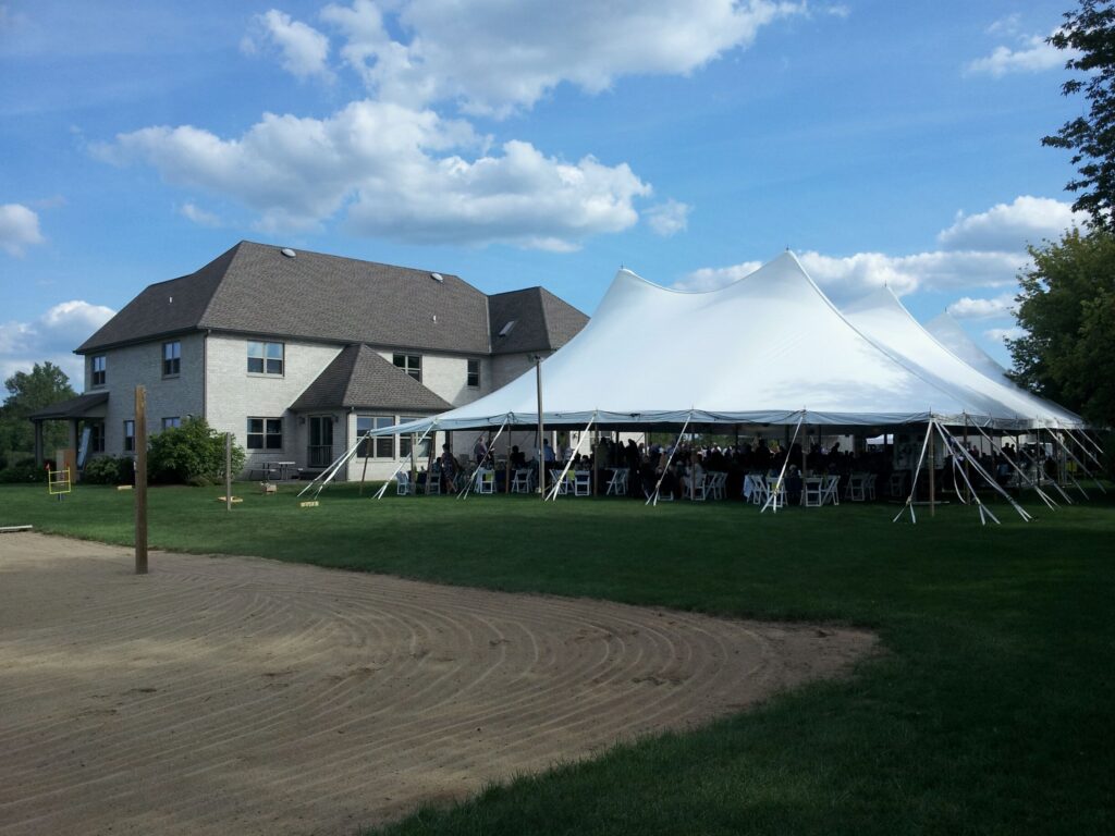 A large white event tent from graduation party rentals is set up on a green lawn beside a two-story house, with people gathered underneath on a sunny day with scattered clouds. Some chairs and tables are visible inside the tent.
