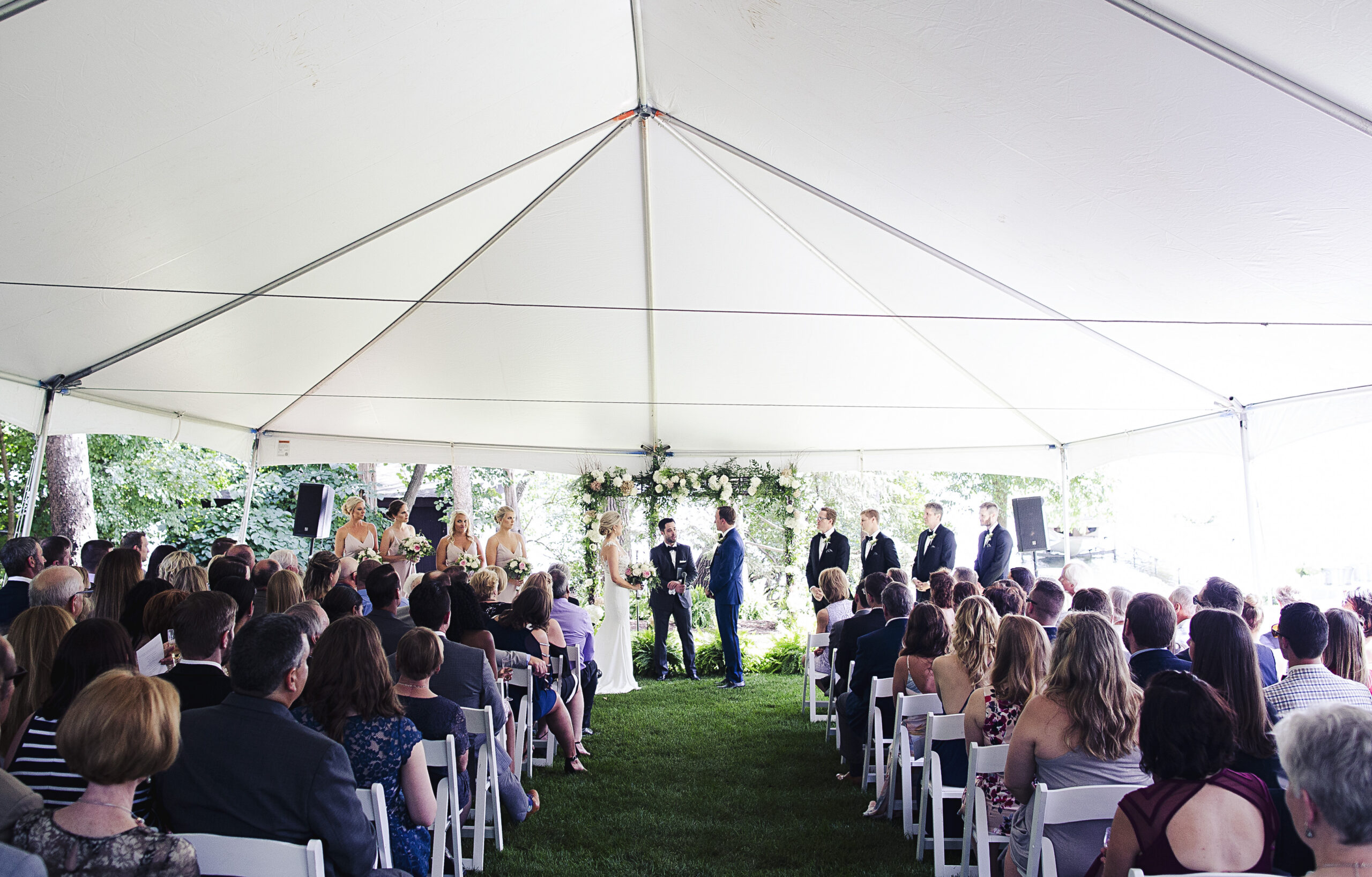 A wedding ceremony takes place under a large white tent, reflecting 2026 wedding rental trends, with guests seated in rows facing the bride, groom, and wedding party at the front, all surrounded by greenery and natural light.