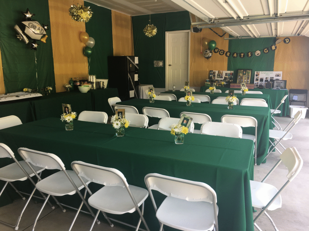 A garage is decorated for a graduation party with green and gold colors. Rectangular tables from graduation party rentals, green tablecloths, white chairs, and flower centerpieces fill the space. Balloons, banners, and photos are displayed on the walls.